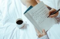 a girl is writing in a notebook sitting with a cup of coffee on a bed